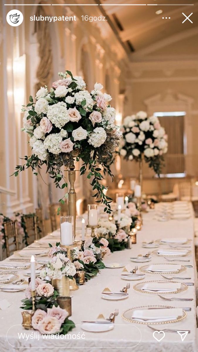 a long table with white and pink flowers on it, surrounded by gold place settings