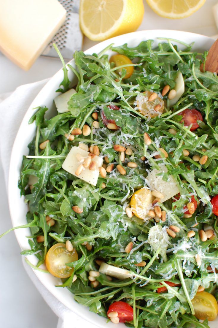 a salad in a white bowl with lemons, tomatoes and pine nuts on the side