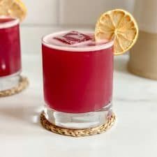 two glasses filled with red liquid sitting on top of a table next to lemons