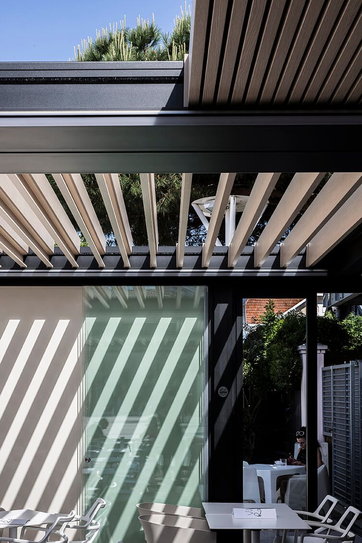 an outdoor dining area with wooden slatted roof and white chairs on the patio