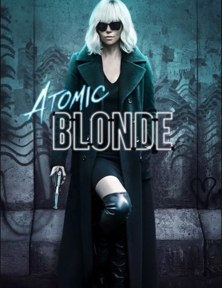 a woman in black coat and sunglasses standing next to a wall with the words atomic blonde on it