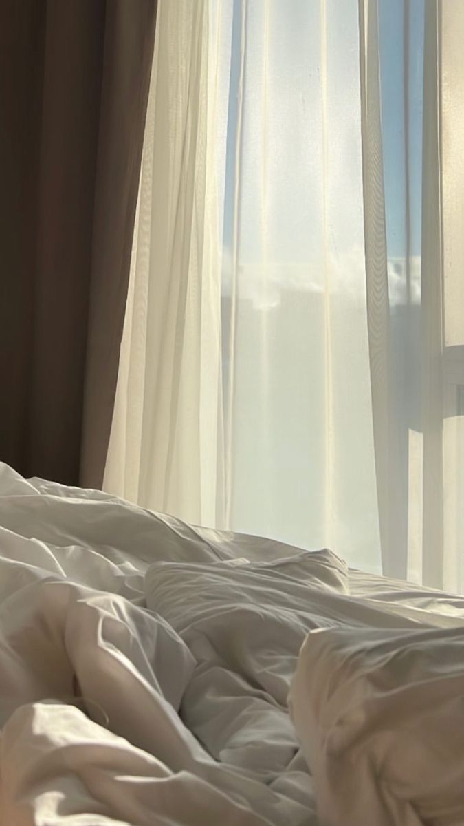 an unmade bed in front of a window with white sheets and drapes on it