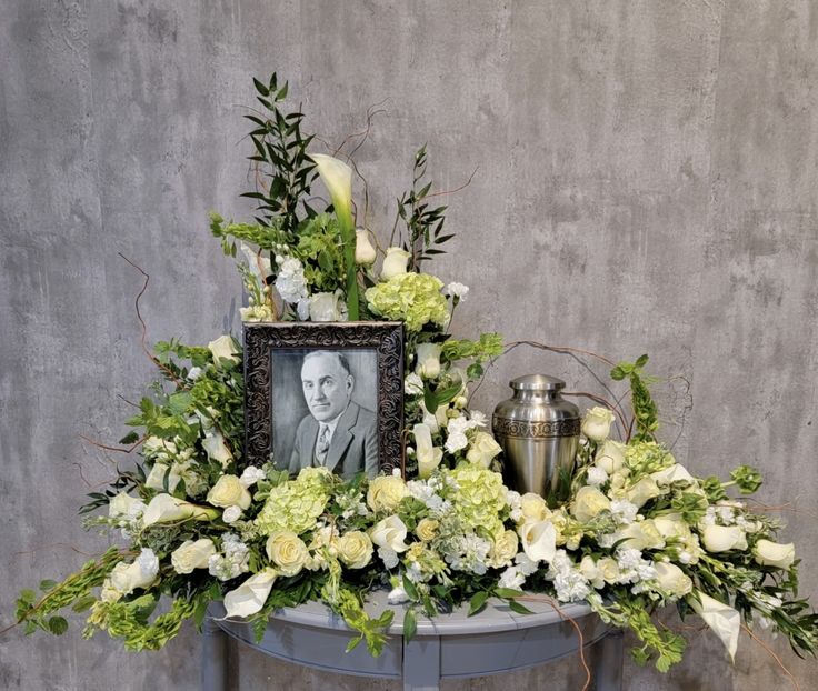 an arrangement of white flowers and greenery on a table with a photo frame in the center