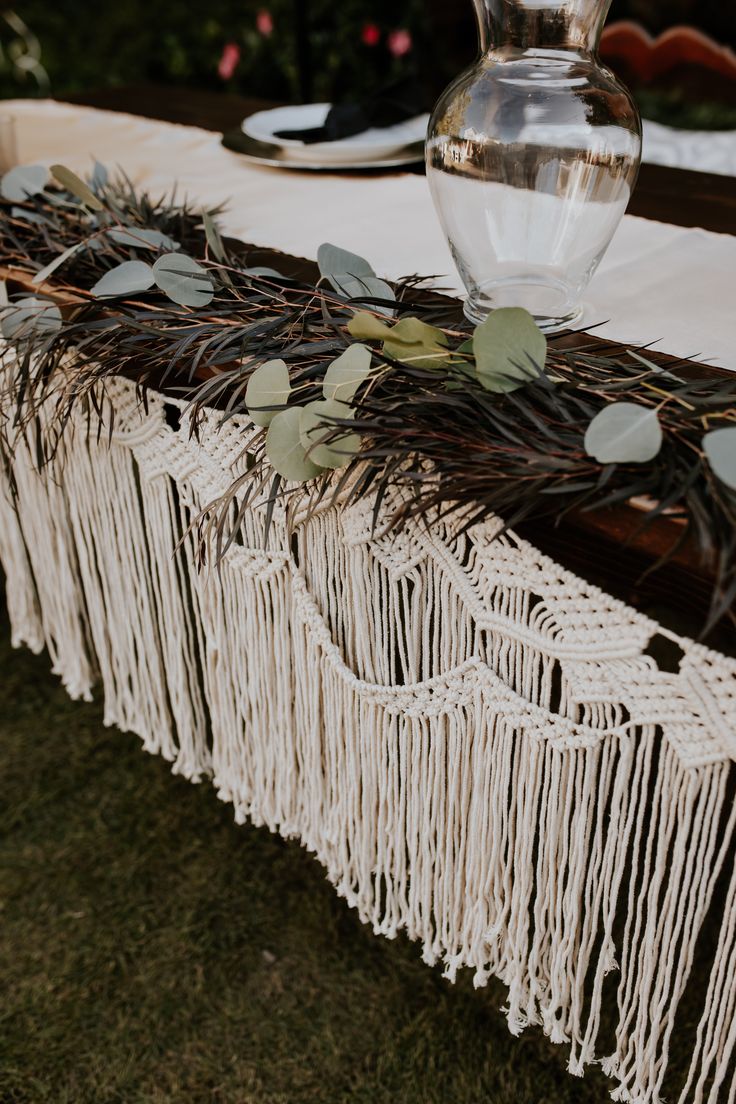 a table topped with a vase filled with water and greenery next to a white table cloth