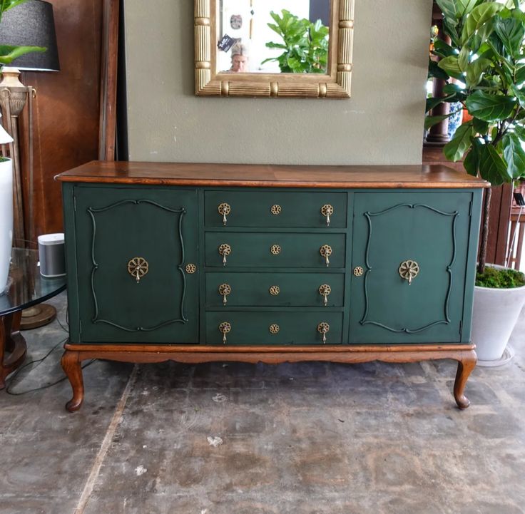 a green dresser with gold knobs on it in front of a mirror and potted plants