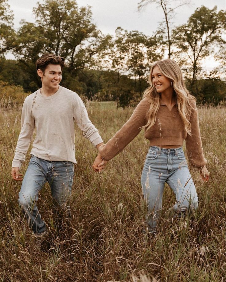 a man and woman holding hands walking through tall grass in a field with trees behind them