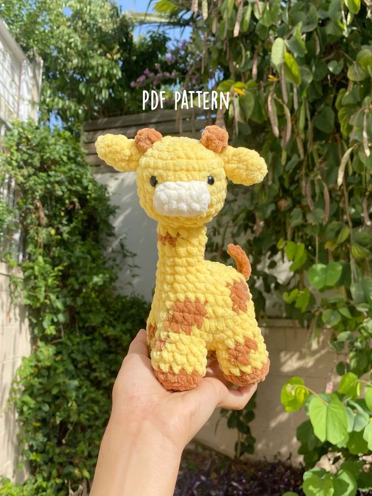 a hand holding up a crocheted giraffe toy in front of some bushes