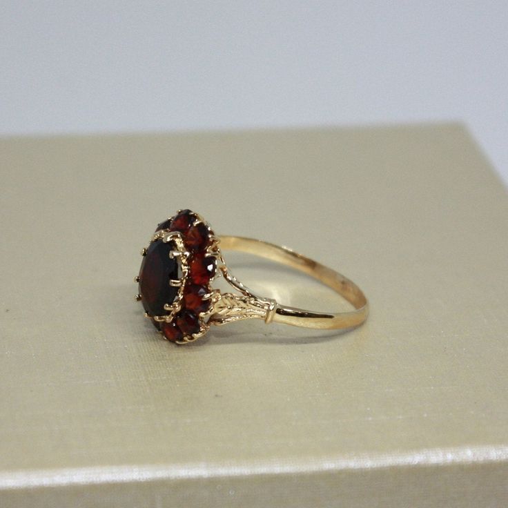 Vintage, Beautiful estate, handcrafted Brown Garnet ring for women, made from 18k solid Yellow gold, Oval shaped natural Garnet in a center surrounded by 10 round smaller stones (Garnets), set on prongs. Total weight of the item is 4g. Polished finish. Excellent condition. Please look at all pictures and If you have any questions, ask, we will be happy to assist you. Note: Store certificate is available, please ask. Item will come in a nice gift box, that will make it look even more special for a someone you love. Item priced for sale. Thank you for shopping with us. Georgian Era Engagement Ring, Red Engagement Ring Unique, 14k Gold Birthstone Ring With Halo Design, Formal Garnet Ring With Center Stone, Yellow Gold Garnet Cluster Ring As Gift, Formal Garnet Rings Fine Jewelry, Formal Garnet Rings With Accent Stones, Yellow Gold Dome Ring With Center Stone, 14k Gold Hallmarked Ring With Round Stone