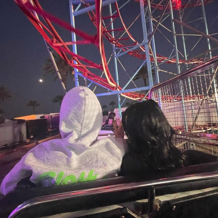 two people sitting in the back of a car at an amusement park, one person wearing a white hoodie