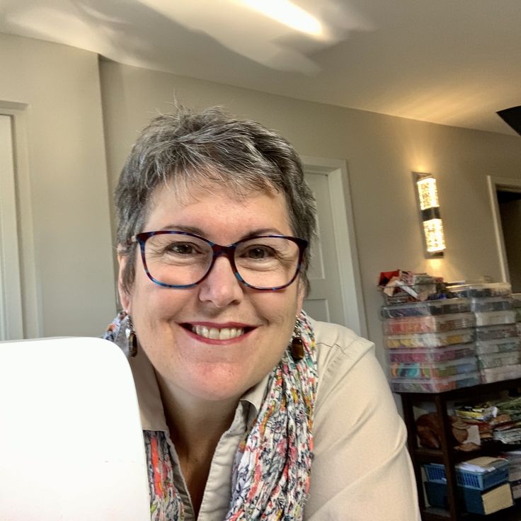 a woman with glasses and a scarf around her neck smiles at the camera while sitting in front of a laptop computer