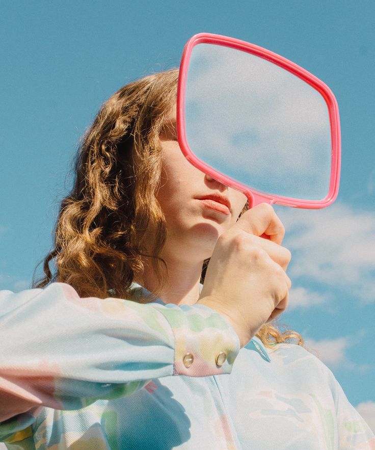 a woman holding a pink mirror to her face while looking at the sky in front of her