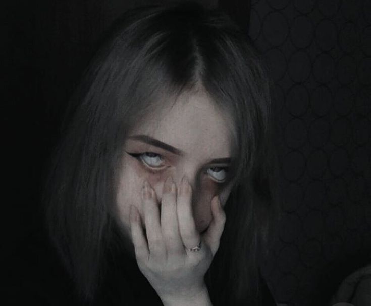 a woman covers her face with her hands while sitting in a dark room and looking at the camera