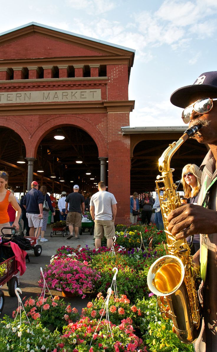 a man playing the saxophone in front of a flower garden with people walking around it