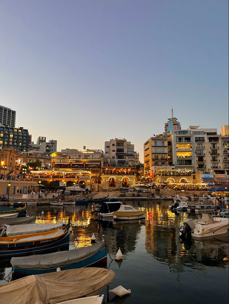 many boats are docked in the water near some buildings and lights at night, with city lights reflected in the water