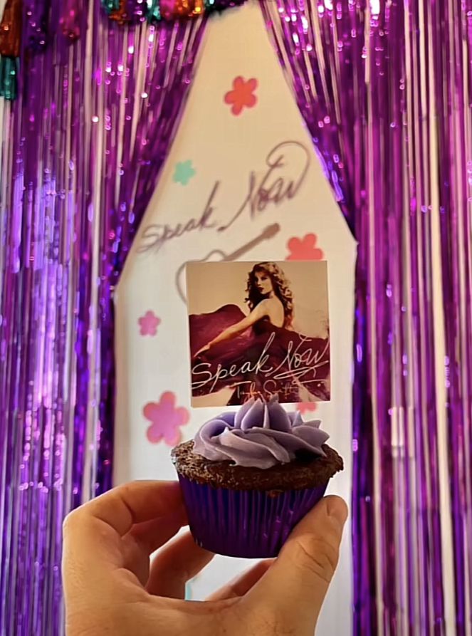 a person holding up a cupcake with purple frosting and a card on top