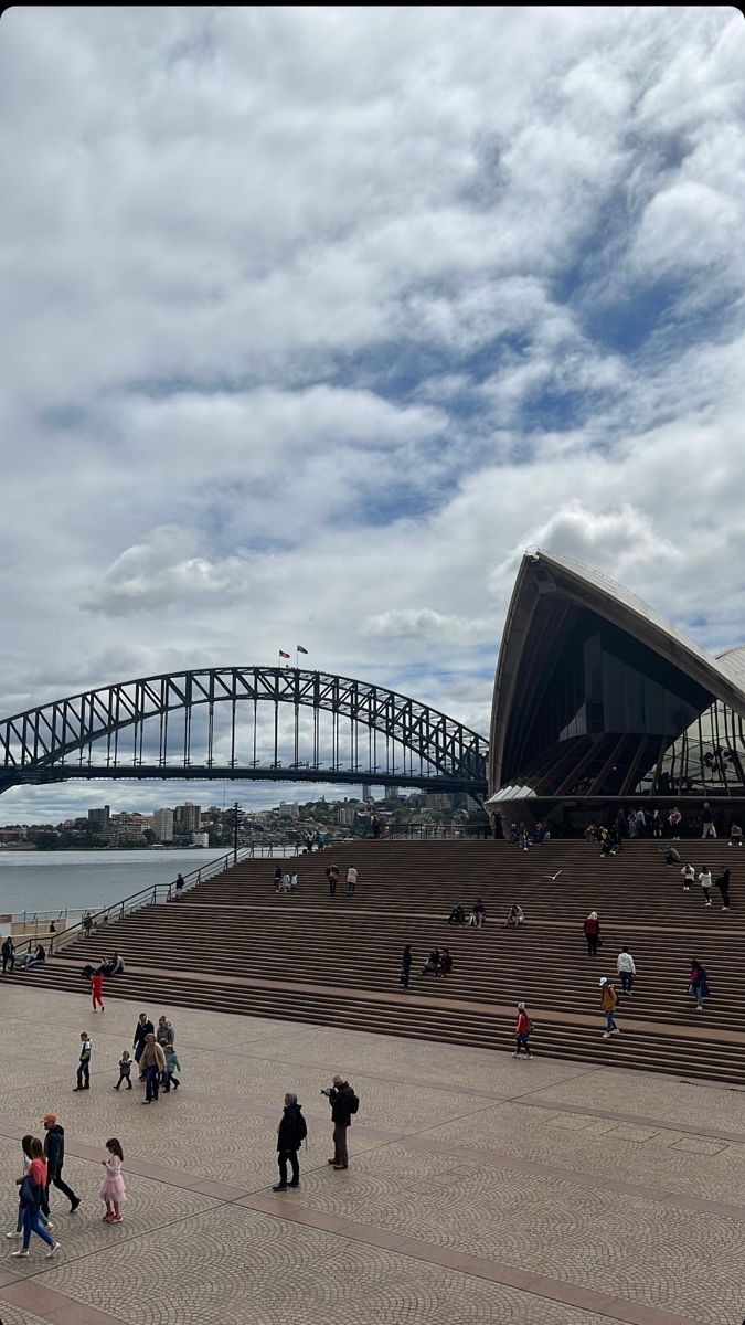 many people are walking around in front of the sydney opera house and harbour bridge on a cloudy day