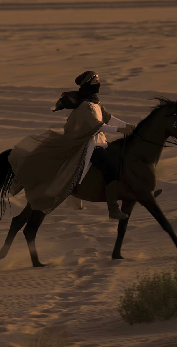 a man riding on the back of a brown horse across a sandy field at sunset