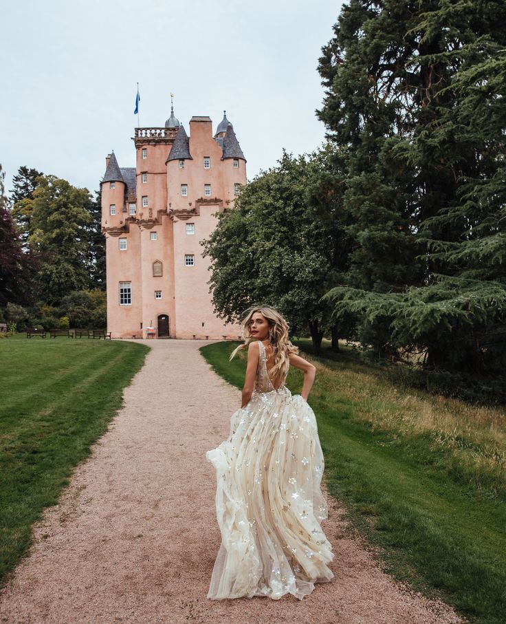 a woman standing in front of a large pink castle with trees around her and wearing a long dress