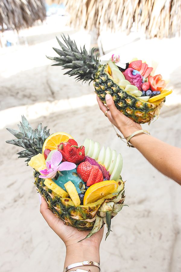 two people holding up baskets filled with fruit on top of a sandy beach next to the ocean