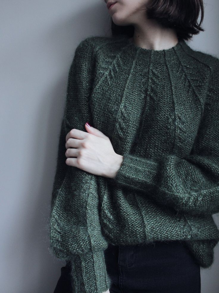 a woman wearing a green sweater standing next to a wall with her hands on her chest