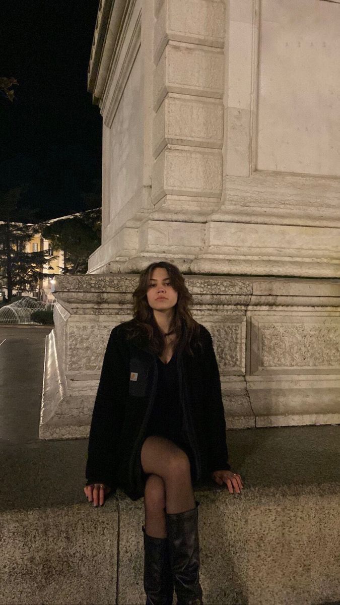 a woman sitting on the side of a wall next to a tall white building at night