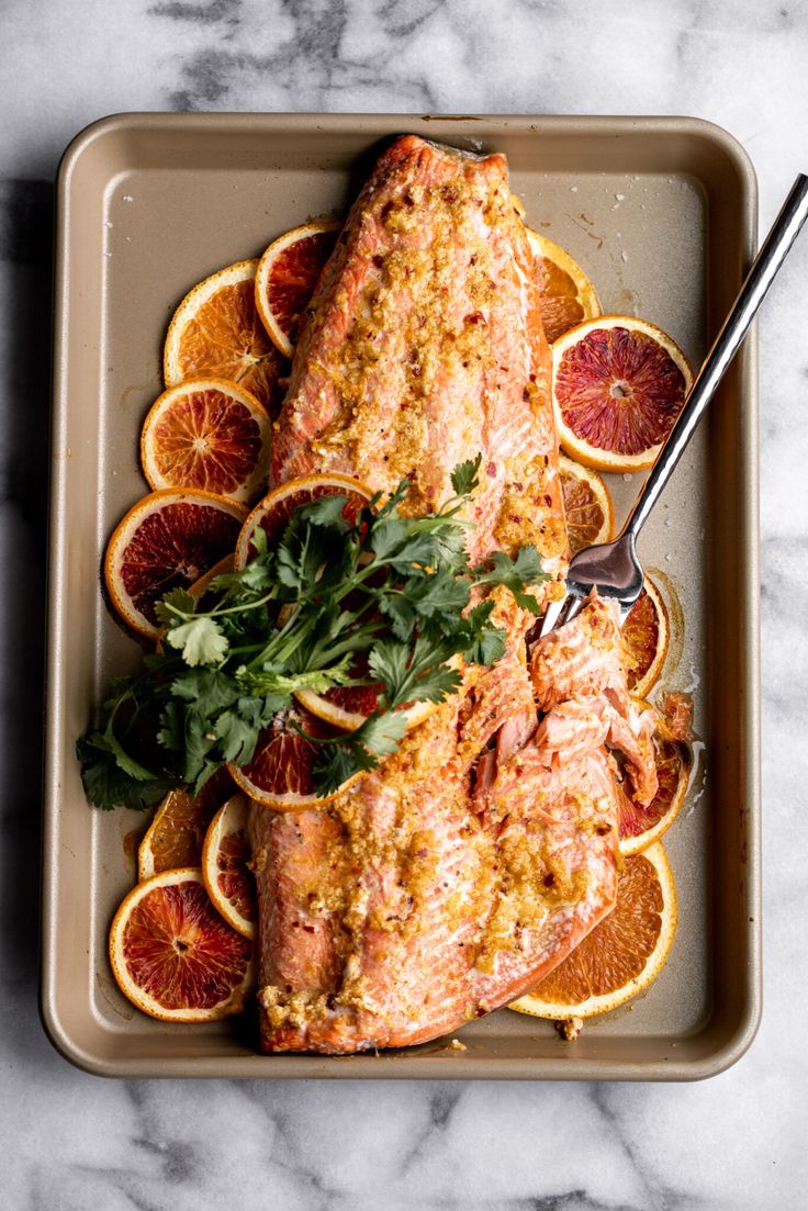 a pan filled with fish and oranges on top of a marble countertop next to a fork