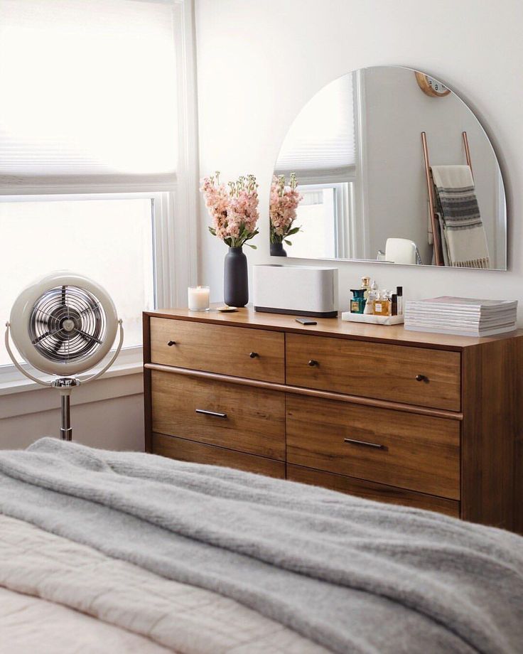 a bedroom with a dresser, mirror and fan in it's corner next to a window