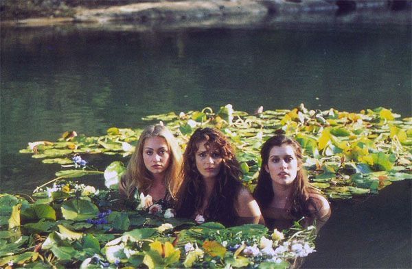 three women are floating in the water surrounded by lily pads and flowers, while one looks at the camera