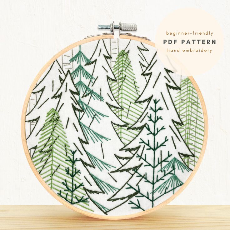 an embroidery pattern with green pine trees in the background on a wooden table next to a white wall