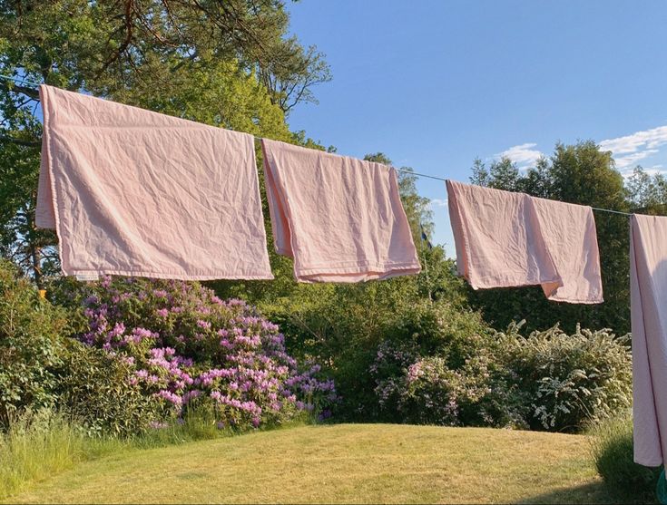 three pink towels hanging on a clothes line in front of some bushes and purple flowers