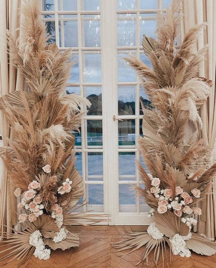 two tall vases with flowers and feathers in front of a window on a wooden floor