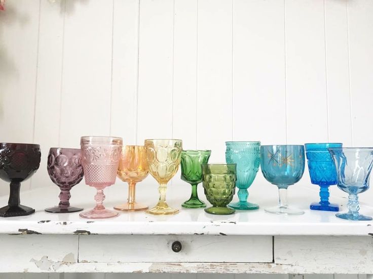 many different colored wine glasses lined up on a shelf in front of a white wall