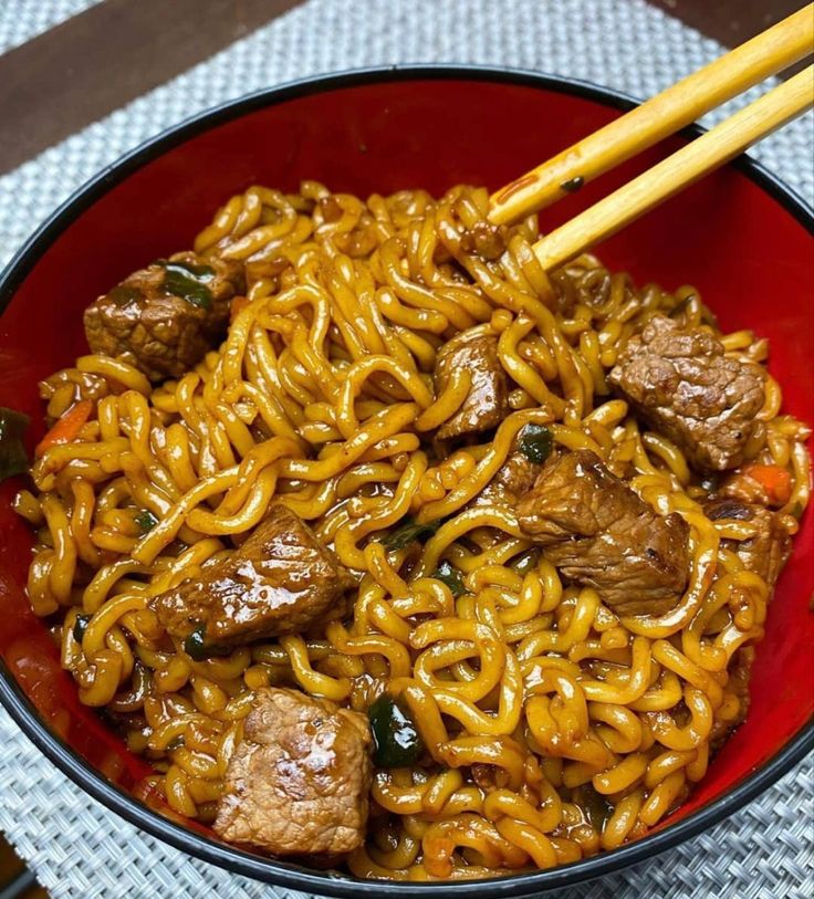 a red bowl filled with noodles and meat next to chopsticks on a table