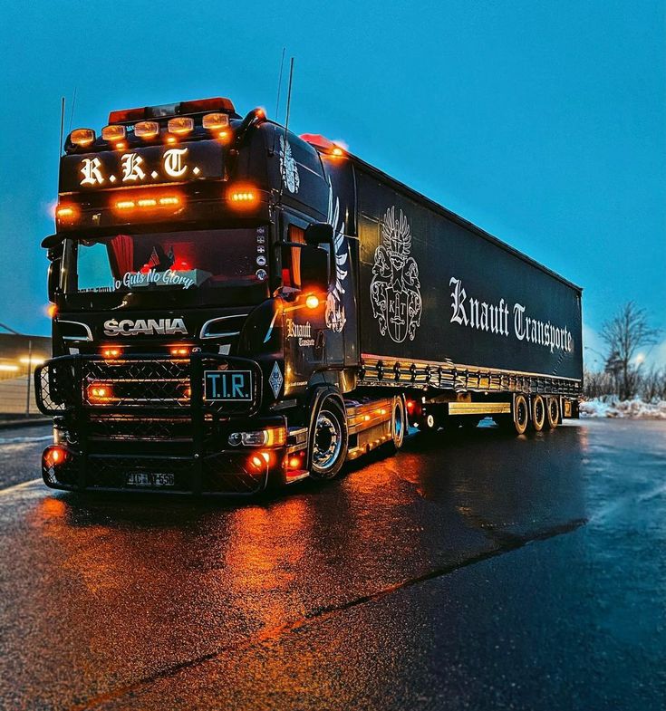 a large semi truck driving down a wet road at night with lights on it's cab