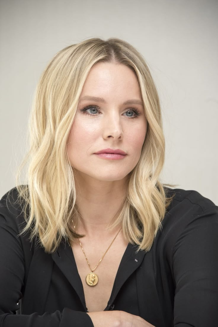 a woman with blonde hair wearing a black shirt and gold necklace sitting in front of a white background