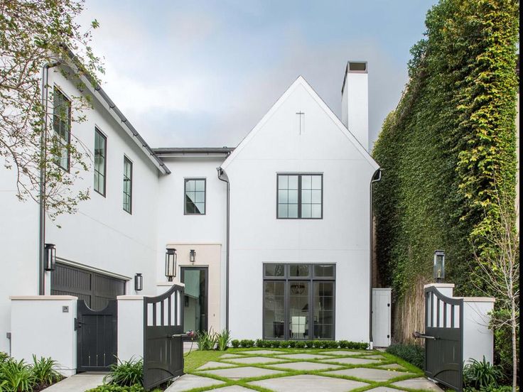 a white house with black gate and green plants on the front lawn, surrounded by greenery