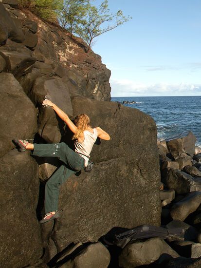 a woman climbing up the side of a large rock next to the ocean on a sunny day