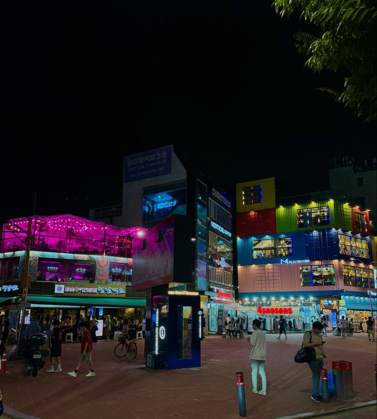 people are walking around in front of a brightly lit shopping center at night with bright lights on the buildings