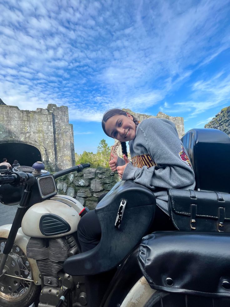 a woman sitting on the back of a motorcycle next to a stone wall with a castle in the background