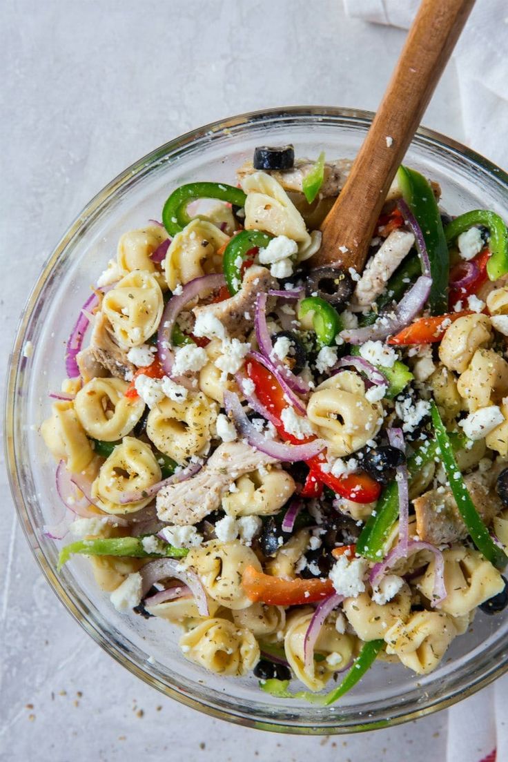 a glass bowl filled with pasta salad on top of a white table cloth next to a wooden spoon