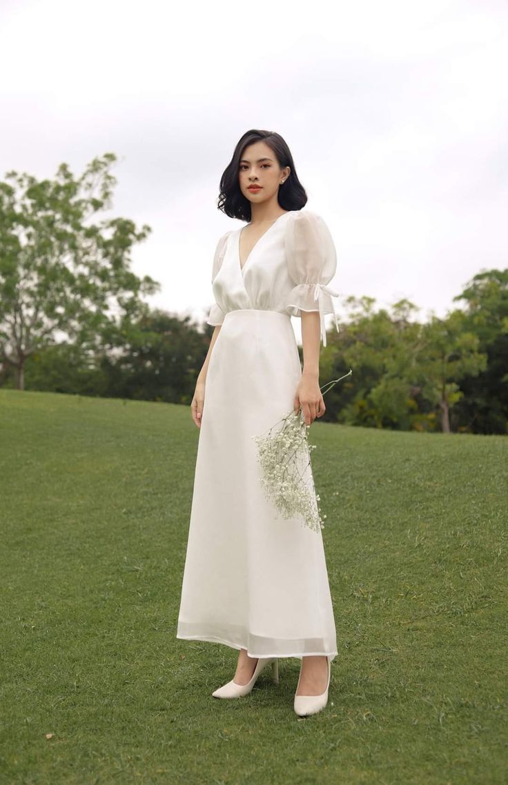 a woman standing in the grass wearing a white dress and holding a bouquet of flowers