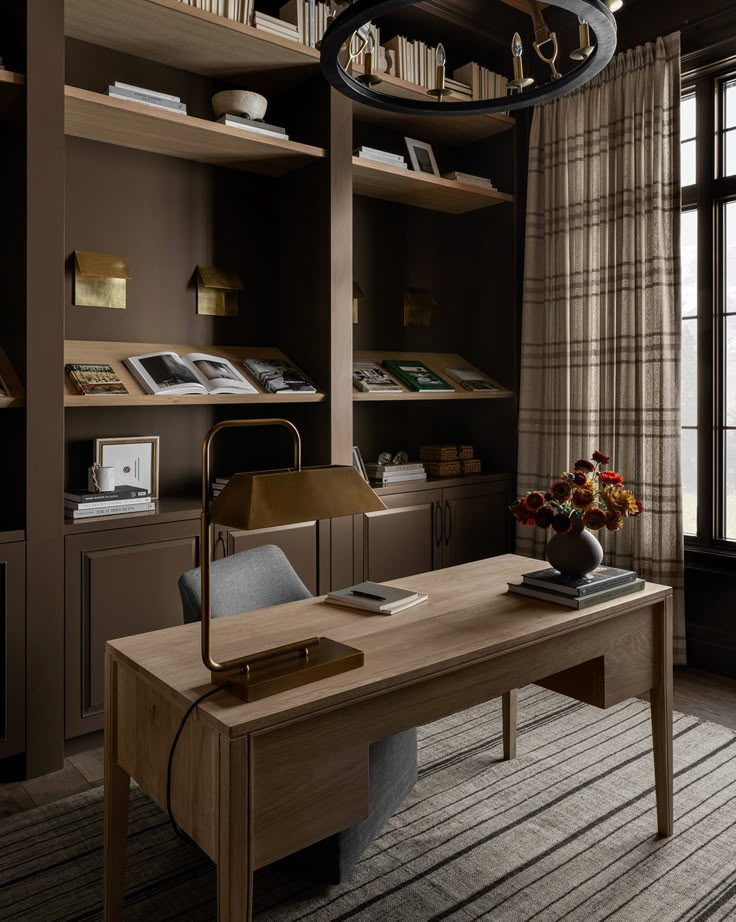 a desk with a lamp, books and flowers on it in front of a window