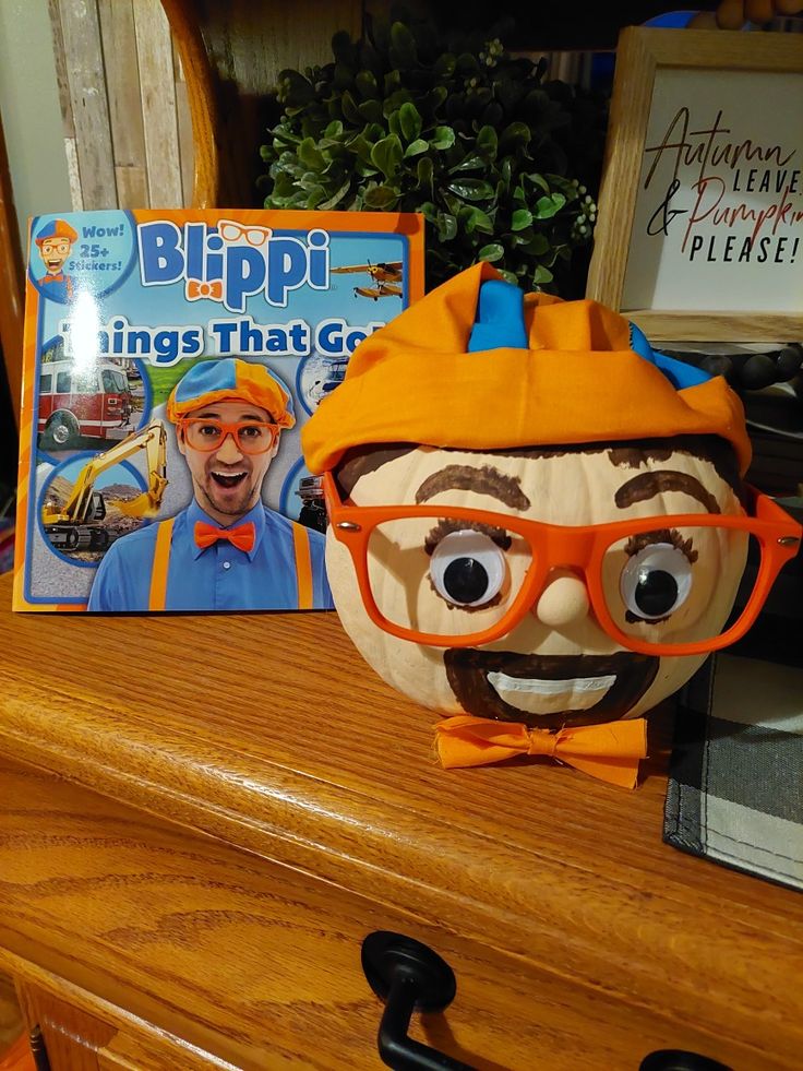 a pumpkin with an orange hat and glasses sitting on top of a wooden dresser next to a book