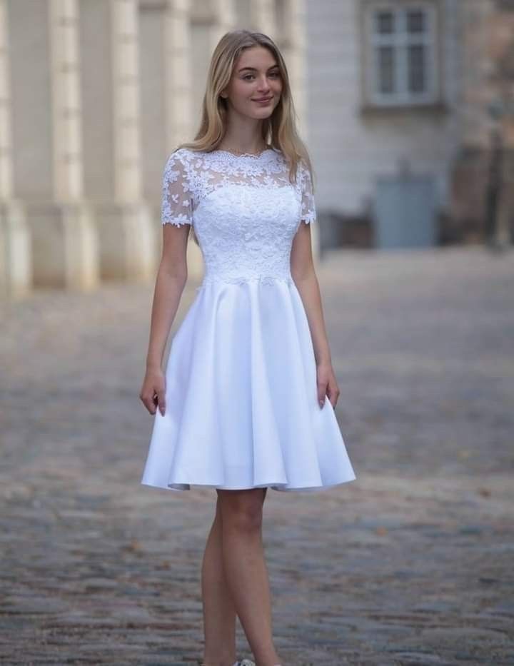 a woman standing in front of a building wearing a white dress with lace on the top