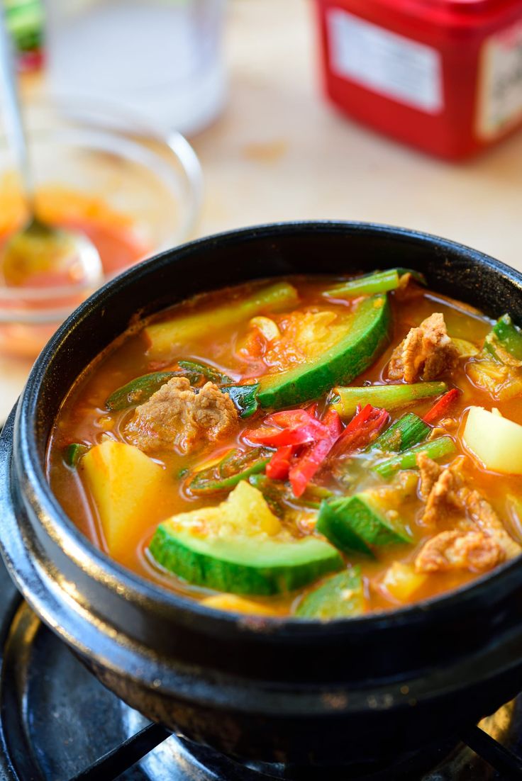 a bowl of soup with meat, vegetables and peppers on the stove top next to a container of seasoning