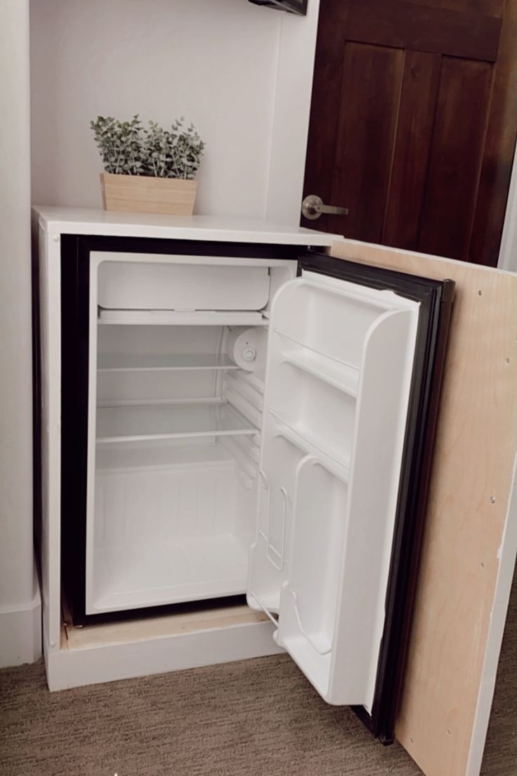 an empty refrigerator with the door open in a small room next to a potted plant