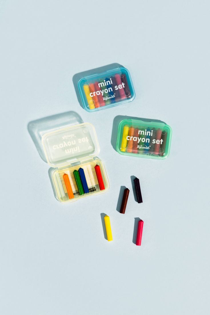 three small crayons in plastic containers on a white surface, one with colored crayons