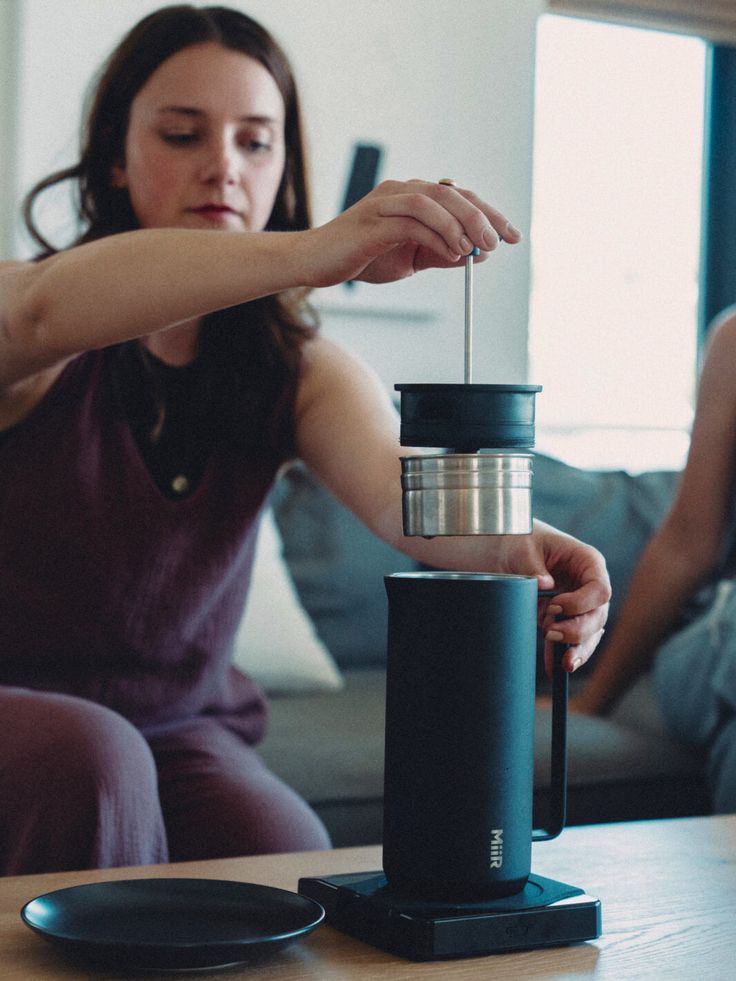 a woman sitting on a couch holding a coffee cup in front of her while another woman looks at the camera