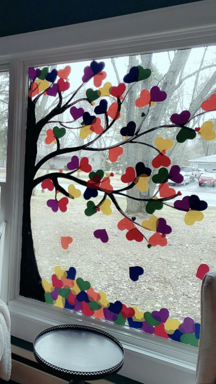 a window with colorful paper hearts hanging from it's side and a tree outside