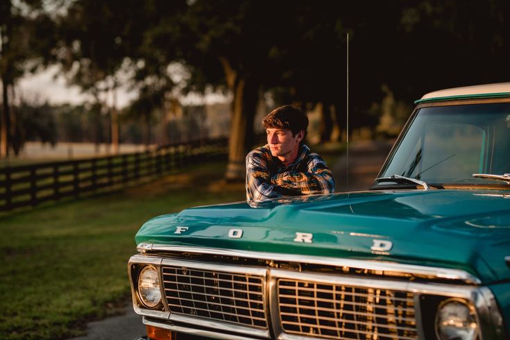 a young man leaning on the hood of an old green ford truck in a field